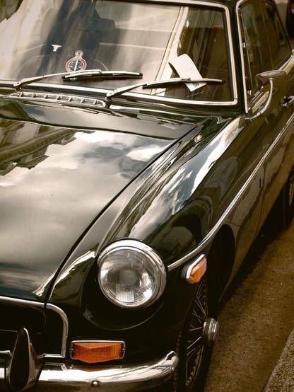 The image shows a classic car parked on a street. It appears to be a vintage model with a dark green color. There's a parking ticket tucked under one of the windshield wipers. The car has a chrome bumper, circular headlight, and classic styling typical of older models. The reflection of buildings and the sky can be seen on the car's surface.