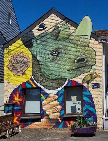 Streetartwall. An imaginative mural with a colorful rhinoceros was sprayed/painted on a single-story exterior wall with two windows. The rhino is wearing a colorful T-shirt and holding a rose. It is a human hand with a tattooed anchor on the index finger. A small black hat hovers above his head. On his shoulder sits a green bird with an implied crown over its head. (There is a large flower pot and a bench in front of the mural.) As strange as the description may sound, the cool rhino man brings a smile to everyone's face.