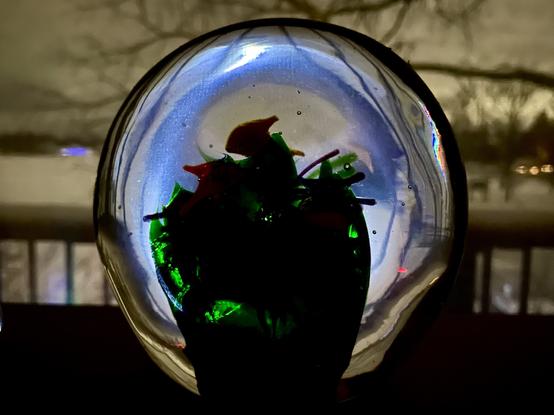 Photograph of a round glass paperweight backlit at night by a window in the background with a porch rail and branches in silhouette behind.