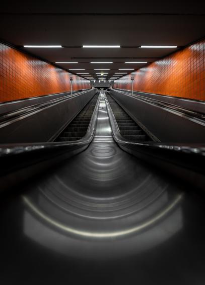 The image shows a symmetrical view of an escalator in an indoor setting, photographed from the base looking upward. The escalator consists of two moving stairways on either side with a stationary, polished metal handrail in the center, which reflects light in circular patterns. The walls on both sides are covered with orange square tiles. Overhead, there are evenly spaced rectangular fluorescent lights embedded in a flat white ceiling, providing illumination. At the top of the escalator, a flat platform with a visible exit sign can be seen. The perspective creates a vanishing point effect toward the center of the image.