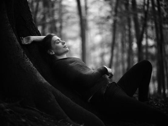 Junge Frau mit einem großen Valpeau-Schultergips, der ihren Oberkörper umschließt, liegt angelehnt an einem Baum in einem Wald und hat die Augen geschlossen.