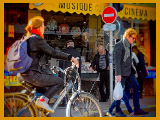 The image shows a street scene with a woman riding a bicycle in the foreground. In the background, there are people browsing outside a music and cinema store. A "No Entry" road sign is visible, and the store has a yellow awning with the words "Musique CD" and "Cinéma" displayed.