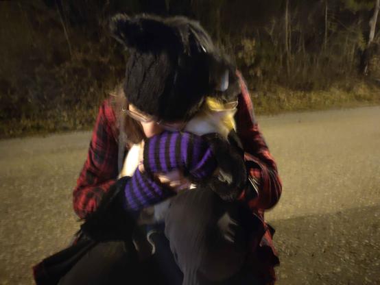 A girl (me) in a red and black coat with a black beanie and cat ears kneeling down on a dimmly lit street. On my lap there's a white and orange colored cat, slightly blurry. The girl has her arms and hands around the cat and holds it thighly, her face leans also down towards the cat.