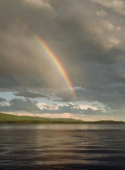 A rainbow streaks across a sky with ominous clouds, over a serene lake with a forest and hillside in the background.