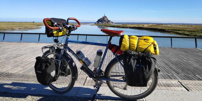 Vélo bleu (Surly Disk Tucker) de voyage équipé avec des sacoches noires et 1 jaune avec guidon papillon et selle marron.
Sur un ponton en bois, au-dessus d'un cours d'eau avec, dans le fond, le Mont Saint-Michel sur un beau ciel bleu.