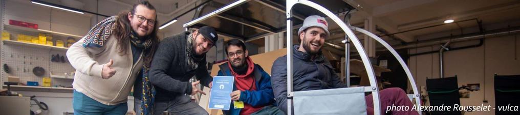 Photo dans un fablab où on voit un vhéliotech (vélo cargo avec toit photovoltaïque) en gros plan. Deux personnes sont installées dessus, l'un d'elle tient un petit panneau au format A4 qui est la certification DIN SPEC 3105 du vhéliotech (mais on ne peut pas lire à cette distance). Deux autres personnes se tiennent à côté. Tout le monde est très souriant.