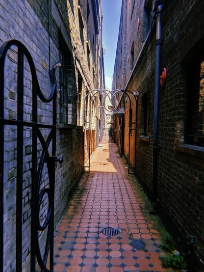 A black metal gate leads into a sunlit red bricked passage way which has more ornate black metal gates further down