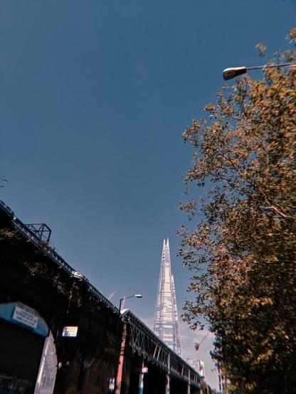 This photo is mostly a bright blue sky. Towering into the sky is London landmark The Shard. This is also a lush green tree and some brick railway arches