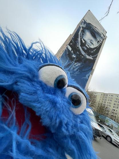 Eine blaue, flauschige Handpuppe mit großen Augen steht im Vordergrund und lächelt in die Kamera. Im Hintergrund zeigt ein hohes Gebäude ein detailliertes Wandbild des Kosmonauten Juri Gagarin.