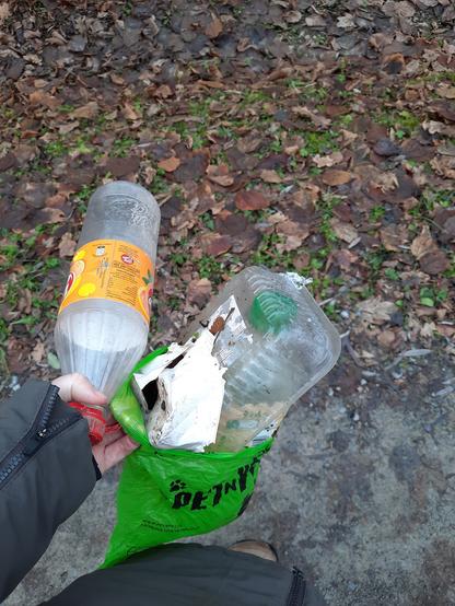 Meine Hand hält eine leere Limoflasche und einen Hundekackbeutel (in dem noch eine Flasche und zwei Kunststoffschalen stecken) ins Bild. 
Im Hintergrund matschiger Feldweg und Laub, mein Ärmel und ein Teil meines Mantels.