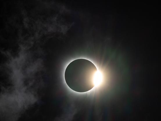 A photo of the total solar eclipse showing the "diamond ring" phenomenon. At the end of totality the sun begins to appear as a bright spot on the right side of the moon, creating the appearance of a shining jewel on a diamond ring. Some clouds are also visible in the sky to the left of the eclipse.