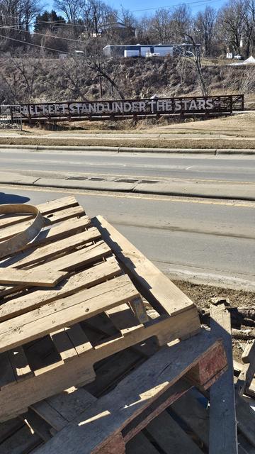 In the foreground, a stack of pallets. Then a street and beyond a long metal sign saying 'All feet stand under the stars'