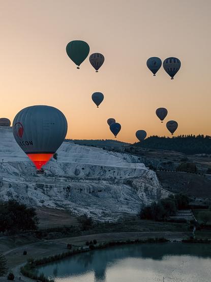 A sunrise in Pamukkale, Turkey, with hot air balloons floating over white travertine terraces. One balloon emits an orange light, and a nearby pond reflects the scene
