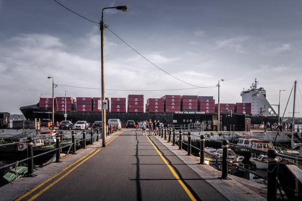 Harbour scene in Cobh showing a large container ship loaded with red containers, with "SPIKE ISLAND TOURS" visible on the dock. A pier walkway leads toward the ship, lined with bollards and small boats moored on either side, under an overcast sky with street lamps overhead.
