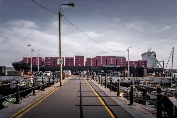 Harbour scene in Cobh showing a large container ship loaded with red containers, with "SPIKE ISLAND TOURS" visible on the dock. A pier walkway leads toward the ship, lined with bollards and small boats moored on either side, under an overcast sky with street lamps overhead.