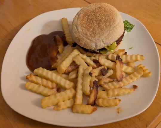 Ein weißer tiefer Teller auf einem Holztisch belegt mit Wellenschnitt Pommes und einem selbst gemachtem Burger. Dazu ein Klecks Ketchup.