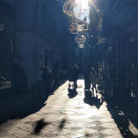 Vista de la calle Zacatín a contra luz. Se ven varias personas paseando. Aún están puestas las luces de navidad. La luz de invierno se refleja en estás luces y crea unas sombras muy alargadas. Los edificios están en sombra y un rayo de luz se desliza por el pavimento.