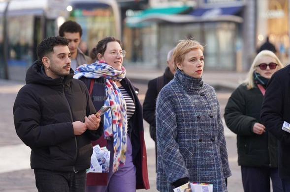 plusieurs personnes debout, en train de marcher vers la droite. De gauche à droite on reconnaît Lyes Louffok, Cyrielle Châtelain et Clémentine Autain. D'autres personnes sont en partie masquées ou hors cadre. En fonds (flou), on distingue un tramway et un décor urbain.