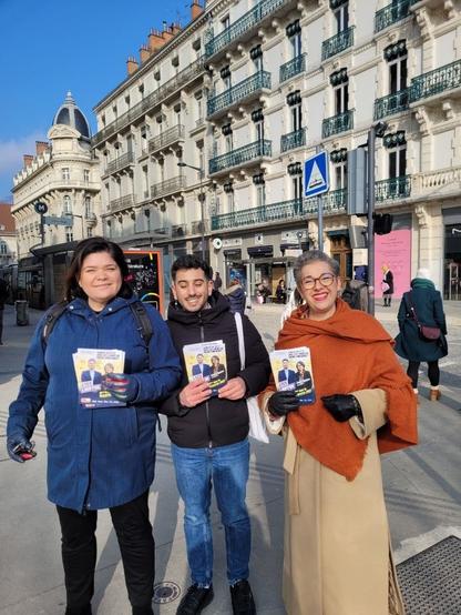 Trois personnes debout de face montrent un tract sur une place. De gauche à droite Raquel Garrido, Lyes Louffok et une militante que votre serviteur ne reconnaît pas, vêtue d'un long vêtement beige et d'un châle couleur brique, cheveux gris, souriante.