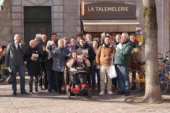 Un groupe d'une vingtaine de personnes, de face, posent avec des tracts. L'un d'entre eux, au premier rang, est en fauteuil roulant. À l'arrière on distingue un local commercial, "La Talemelerie"