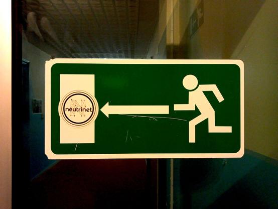 Photo d'un panneau indiquant le chemin vers une issue en cas d’incendie avec un personnage qui court, une flèche et une porte. Le personnage court vers la porte et la flèche lui indique le chemin. Sur la porte, un sticker de Neutrinet a été collé.