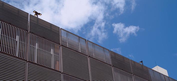 Photograph looking up at a multi-storey car park.  The side of the carpark is made of square patches of steel strips. Each patch has strips running vertically or horizontally, making a chequerboard pattern on the side of the building.  A light pole is peeking above the top floor.
Blue sky with a few scattered clouds is visible above the building.