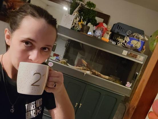 Me, Kiwi, drinking out of a white mug with "21" written on the front in gold, stood in front of a large grey wooden, glass fronted enclosure.