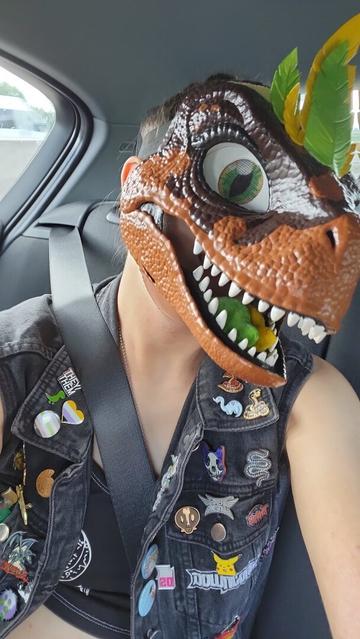 Me sat in the back of a car wearing Coffee, my brown dinosaur mask with yellow and green feathers, and a black non-sleeved denim jacket that is heavily decorated with metal pins.
