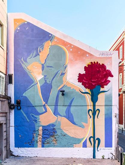 Streetartwall. A beautiful mural of two people and a rose is painted on the outside wall of a single-storey building. It shows the figures in delicate pastel shades. From behind, a young man (or woman) with half-length hair can be seen holding and kissing the head of a young bald man. The kissed person's hand grasps the arm of the other. An orange starry sky can be seen in the background and the left side is taken up by a large painted red rose in bold colors. An unusual but beautiful composition. 
Info: According to the artist, the mural is about "... Freedom that only comes from the search for knowledge and information. Freedom comes from equality. Fear, silence, shame and censorship prevent us from sharing the different perspectives of truth and thus hinder our development and growth. Freedom means being aware of our development."