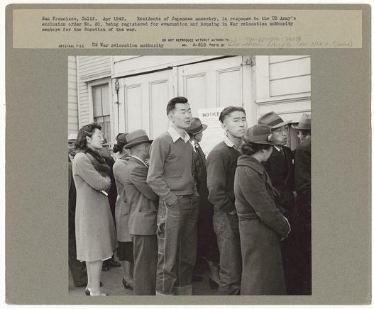 The image depicts a group of individuals, likely from Japanese ancestry, standing in line during the early 1940s. They are outside a building with an announcement about registration for evacuation and housing under America's War Relocation Authority due to Exclusion Order No. 20.

Several people can be seen wearing winter coats, indicating it might be cold weather at that time. Some of them appear focused on the task ahead while others seem contemplative or concerned. A woman in a coat with fur trim stands out among them, and there are men dressed in suits, some donning hats, which is typical for attire during the mid-20th century.

The photograph bears text at the top that reads "San Francisco, Calif., Apr 1942," providing context to the location and date. Additional information includes a reference number (A-518), photographer's name (Dorothea Lange), and notes about the original file being from US War relocation authority.

The overall mood of the image conveys somberness, as these individuals are partaking in what is likely an involuntary process due to wartime anti-Japanese sentiment. The photograph captures a historical moment during World War II when Japanese Americans faced exclusion and forced relocation by American authorities under military orders.