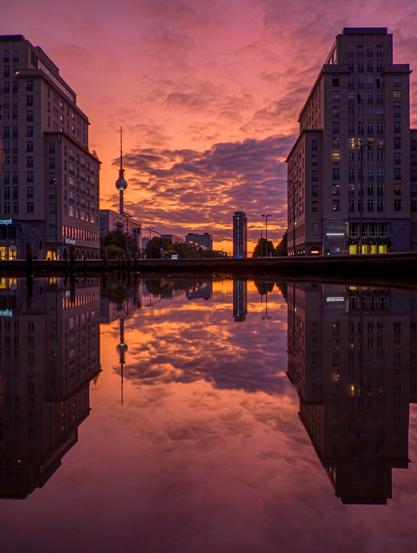 An urban scene featuring two tall buildings on either side of a body of water, symmetrically reflected on the calm surface. In the center of the image, the Berlin TV Tower rises into the sky. The sky displays vibrant hues of orange, pink, and purple, enhanced by the low sun and dense cloud formations. The reflection in the water perfectly mirrors the cityscape, creating a serene yet striking visual composition.