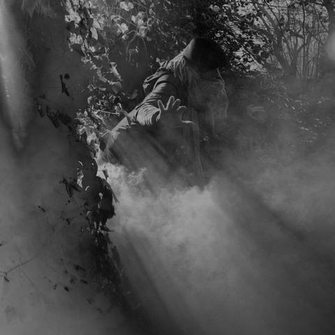 Digital black and white picture of a woman stepping out from between some trees. She is lit from the back. Smoke is visible in the shot, creating a mysterious atmosphere.