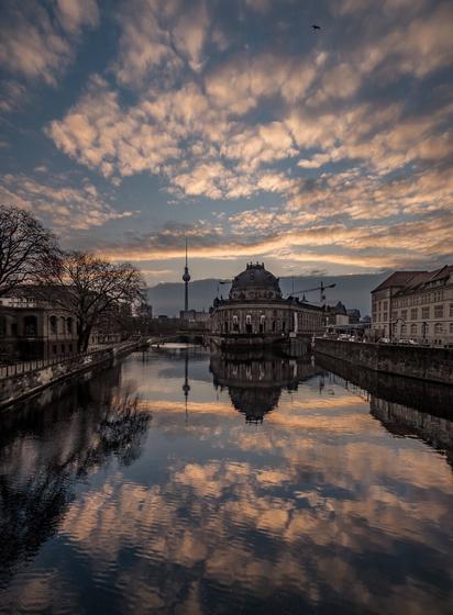 The image depicts an urban scene during sunrise or sunset. In the foreground, a calm river reflects the surroundings and the clouds in the sky clearly. On the right side stands the Bode Museum with its distinctive dome. In the background, the Berlin TV Tower rises above the skyline. Buildings and bare trees line the riverbanks on both sides. The sky is filled with warm orange and blue clouds. A single crow is flying in the upper part of the image.