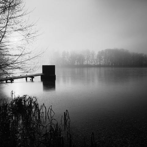 Monochrome image of a frozen lake in a backlight scene. In the left of the image is a lake monk, which looks almost black.
