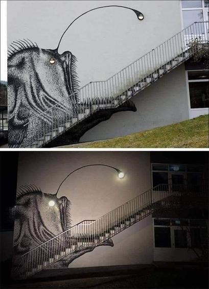 A striking street art mural by SKURK at Rå Skole in Bergen, Norway, creatively incorporates architectural features into its design. The mural depicts an anglerfish, with its body painted along the side of a white building, and a staircase cleverly forming the fish's mouth and teeth. A streetlight at the top of the staircase doubles as the anglerfish's glowing lure. The artwork is equally impactful during the day, showcasing its detailed black-and-white stippling technique, and at night, when the illuminated light transforms the mural into a vivid and dynamic scene. This masterpiece exemplifies the seamless blending of art and architecture.