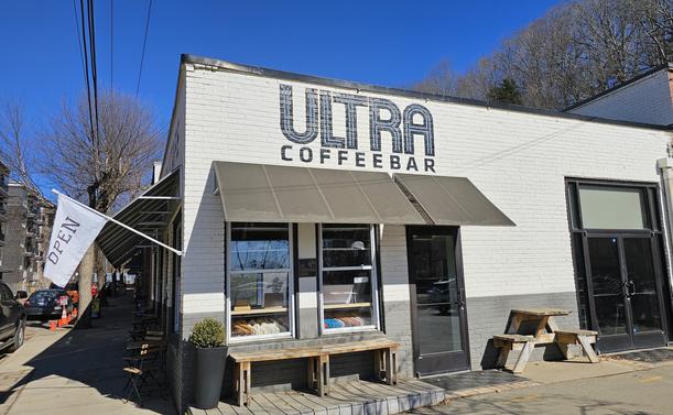 The front of a coffee shop painted white. There is a flag pointing out to the left saying "Open". Above an awning is painted Ultra Coffee Bar. A bench and picnic table sit in front. Leafless trees rise above the rear of the building under a bright blue sky.