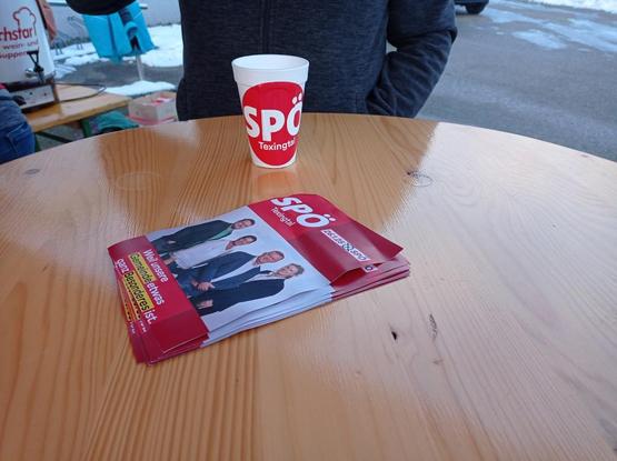 Ein runder Stehtisch aus Holz. Auf dem Tisch steht ein Styroporbecher mit einem Aufkleber und der AUfschrift SPÖ Texingtal. 

Vor dem Becher, in der Mitte des Tisches liegen viele Flyer mit Wahlkampfflyern