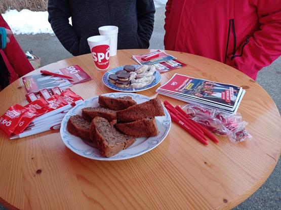 Ein runder Stehtisch aus Holz. Auf dem Tisch stehen ein Styroporbecher mit einem Aufkleber und der Aufschrift SPÖ Texingtal, sowie unterschiedliche Werbemittel wie Kugelschreiber, Gummibärchen und Flyer.  

Vor dem Becher, in der Mitte des Tisches stehen Teller mit Keksen und Kuchen. 

Hinter dem Tisch sind drei Personen in Jacken zu sehen.