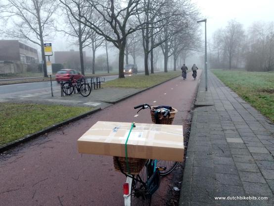Bicycle on a cyclepath with parcels loaded on the back. The cyclepath also has other cyclists on it. There are cars on the road, spaced from us by a large verge, making it's safe to cycle in fog.