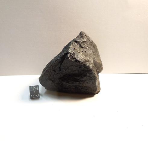 A fist-sized mass of black Zhamanshinite sitting next to a black centimeter scale cube.