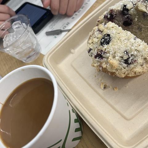 A close-up of a filled paper coffee cup (with cream) next to a large muffin, topped with crumbly streusel and visible blueberries. A clear water cup with ice is nearby, and a hand is seen holding a smartphone in the background.