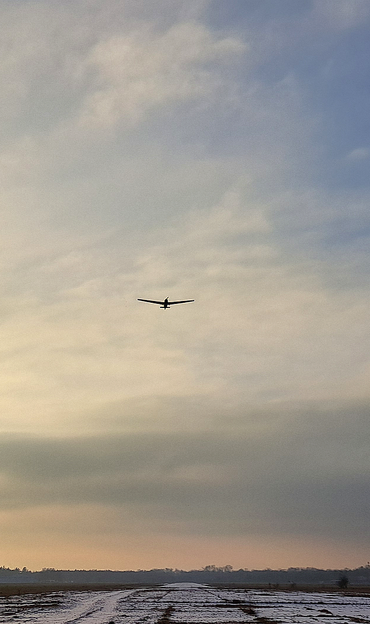 Ein kleines, dunkles Flugzeug fliegt in geringer Höhe über einer leicht verschneiten, flachen Landschaft. Der Himmel ist bedeckt mit zarten, hellen Wolken, die in Pastelltönen von Beige, Rosa und Blau gehalten sind. Am Horizont ist ein dunkler Waldstreifen zu sehen. Das Bild wirkt ruhig und friedlich, mit einem Hauch von Abend- oder Morgenlicht.

Bereitgestellt von @altbot, generiert mit Gemini