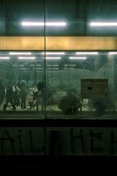 Photo of people waiting for buses or trams at an underground station, taken through a dirty glass. Dim lights above give the scene an eery quality. The tones of the photo are greenish and beige.