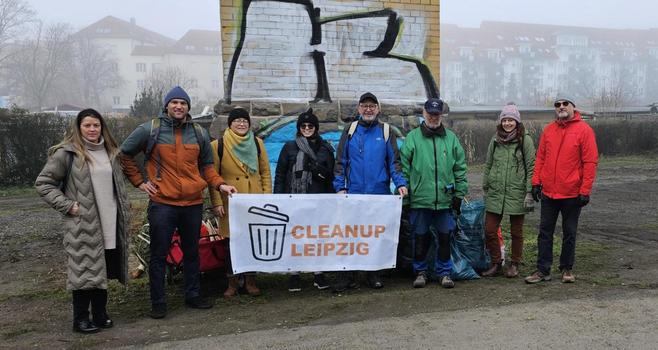 Eine Gruppe von etwa 8 Freiwilligen steht vor einem mit Graffiti besprühten Mauerwerk in 
Leipzig. Sie halten ein Banner mit der Aufschrift "Cleanup Leipzig". Sie tragen warme Winterkleidung und scheinen gerade eine Müllsammelaktion beendet zu haben. Im Hintergrund ist ein nebliger Tag und ein Wohnhauskomplex zu sehen. Das Bild vermittelt ein Gefühl von Gemeinschaftsgefühl und Engagement für Umweltschutz.