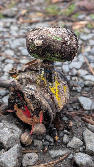 Das Foto zeigt ein verwittertes, altes Laufente aus Holz, das auf dem Boden liegt. Es scheint ein selbstgebautes oder handgefertigtes Spielzeug zu sein, mit einem runden Kopf und einem zylindrischen Körper. Die Oberfläche ist mit Schmutz, Erde und Ablagerungen bedeckt, und Reste von Farbe, in Gelb und Grün, sind noch sichtbar. Kleine, runde Holzstücke sind an Schnüren an der Unterseite befestigt. Das Spielzeug liegt auf einem Untergrund aus kleinen Steinen und Erde.