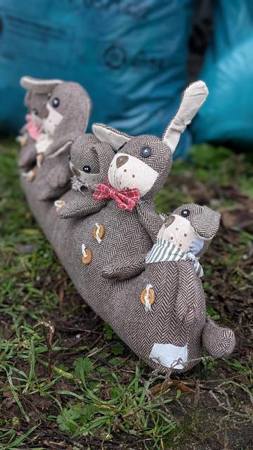 Ein Türstopper in Form einer Reihe von niedlichen Stoffhasen liegt auf dem Boden im Freien. Die Hasen sind in braunem Tweedstoff gehalten und haben große, niedliche Ohren. Sie sind unterschiedlich groß, der größte Hase befindet sich in der Mitte. Jeder Hase hat Holzknöpfe als Dekoration. Der Türstopper liegt auf einer Wiese mit Gras und Erde. Im Hintergrund ist ein blauer Müllsack der Stadtreinigung Leipzig unscharf zu sehen.