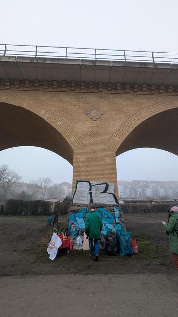Das Foto zeigt eine große, gelbliche Ziegelbrücke mit zwei Bögen, ein Viadukt. Unter der Brücke stehen zwei Personen, die Müll einsammeln. Ein Teil der Brücke ist mit Graffiti besprüht. Im Hintergrund sind Wohnhäuser und Bäume zu sehen. Der Himmel ist bedeckt und es ist ein eher trüber Tag. Die Szene wirkt wie eine Müllsammelaktion in einer städtischen Umgebung.