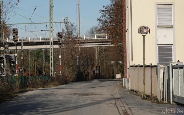 Hochstehendes Lichtsperrsignal in Stellung Hp 0 am Rande einer asphaltierten Straße. Im Hintergrund links zwei Ausfahrsignale hinter einem Zaun. Auf der Straße lassen sich noch die überteerten Reste der Schienen erkennen, die in Richtung der Ausfahrsignale verlaufen.