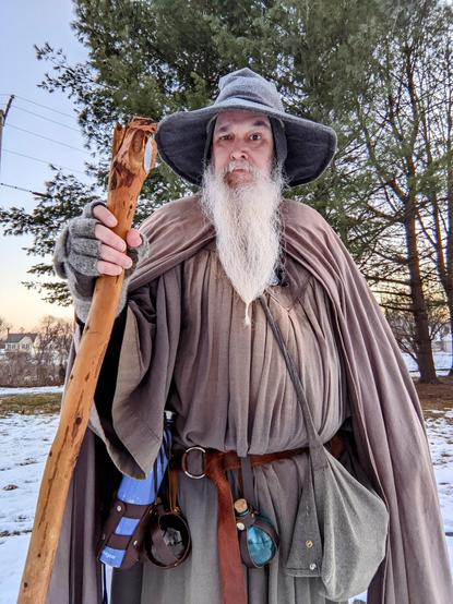 middle aged man with long grey beard and Gandalf style outfit with hat & staff stands outside at dusk in front of a grove of trees raising one eyebrow at the camera