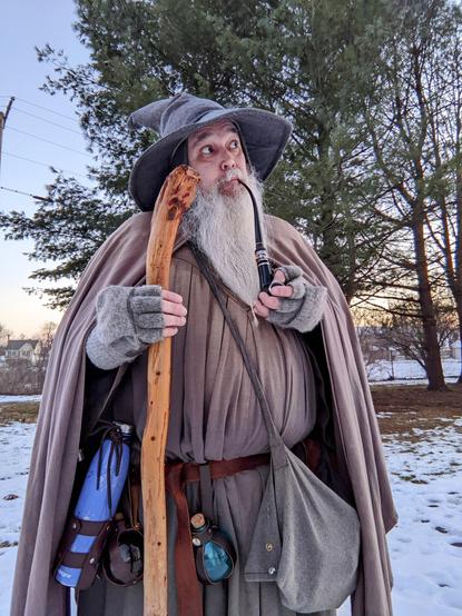 middle aged man with long grey beard and Gandalf style outfit with hat & staff stands outside at dusk in front of a grove of trees with a pipe looking up and to the right with wonder